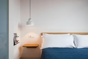 A minimalist bedroom featuring a white bed with a blue blanket and white pillows, under a pendant light and a floating nights