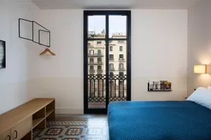 A minimalist bedroom featuring a blue bed with a wooden headboard, white walls, and a large window with a metal railing.