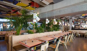A modern industrial restaurant interior featuring concrete floors, wooden furniture, and green plants.