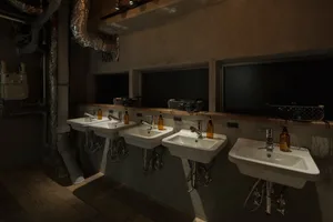 Multiple white basins with orange soap dispensers in a dimly lit, industrial-style public bathroom with concrete walls and ex