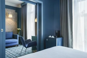A modern hotel bedroom with blue walls, a white bedspread, a blue sofa, and a chair.