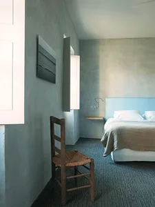 A minimalist bedroom featuring a concrete headboard, white furniture, and a woven chair.