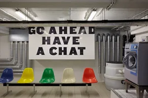 A modern office lounge area with colorful chairs arranged in a row, featuring industrial-style tiles and concrete ceiling.