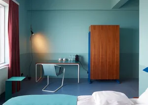 A minimalist bedroom with blue walls, wooden furniture, and a desk with a lamp and coffee mugs.