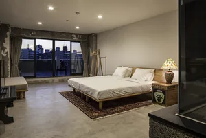 Modern bedroom with concrete floor, wooden bed frame, and rug, featuring a cityscape view from a window