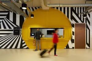A modern hotel lobby featuring a bright yellow metal reception desk with black and white striped walls.