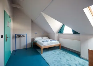 A minimalist bedroom with blue flooring and wooden furniture, featuring a bed with white bedding and a small window.