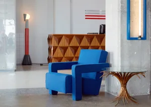 A modern minimalist lobby featuring a blue armchair, concrete floor, and a table made of glass and natural elements.