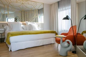 A modern bedroom featuring a bed with a white bedspread and gold metal frame, an orange upholstered armchair, and a side tabl