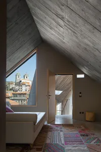A modern living room with concrete walls, a window overlooking a cityscape, a sofa, and a patterned rug.