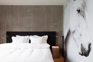 A modern minimalist bedroom features a wall of concrete and white bedding, illuminated by minimalistic lighting.