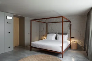 Spacious and minimalist bedroom with exposed concrete walls, a wooden canopy bed, and a circular woven rug.
