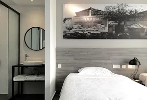 A minimalist hotel room with a single bed, a sink, and a framed black and white photograph of a street scene hanging on the w