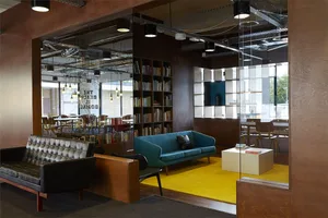 A modern office space featuring metal-framed furniture, with a mix of seating options, a bookshelf, and large windows.