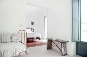 Minimalist bedroom with a white floor, wooden bench, and a bed with white bedding and a single striped cushion.