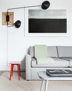 A modern grey sofa with a green throw blanket, white walls, and black floor lamp, accompanied by a red stool.