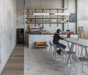 Modern cafe bar with concrete walls, wooden tables, and seating arranged around a central bar counter.