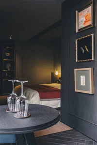 A dark-colored wine glass placed upside-down on a wooden table with a black wood floor and a bed with a red blanket in the ba