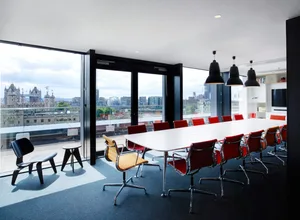 A modern conference room featuring a long white table surrounded by red chairs, black pendant lights, and a view of a citysca