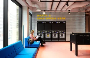 A person sitting on a blue cushion in a modern industrial laundromat lounge with concrete flooring and washing machines in th