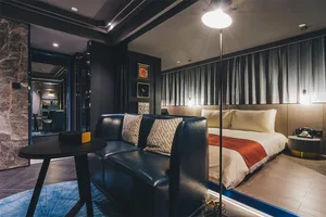 A modern living room with a black leather couch, white bed with red blanket, and dark curtains.