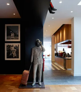 A modern statue of a person dressed in a patterned suit and holding a suitcase stands in a lobby with black and white checker