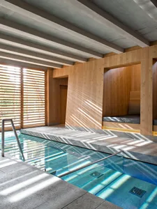 An industrial-style poolside area featuring concrete flooring and wooden seating with natural light streaming through wooden 