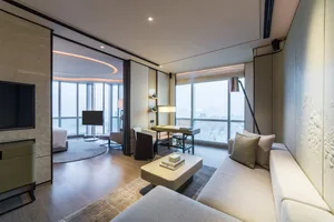 A modern minimalist living room with white decor, featuring a large sofa, coffee table, and desk area with a cityscape view.