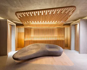 A minimalist lobby with wooden accents and concrete flooring features a large, organic-shaped rock-like object in the center.