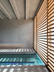 A minimalist indoor swimming pool with concrete walls and light streaming through a wooden slatted wall.