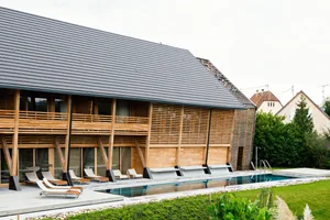 A modern residential exterior with a wooden facade and a pitched roof with a pool in the foreground.