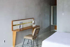 A minimalist bedroom with a concrete headboard and floor, featuring a white bed and a simple desk area.