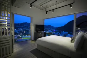 Modern minimalist bedroom with a city view, featuring a large bed, a television, and large windows overlooking the cityscape 