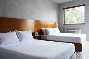 A minimalist bedroom with two double beds, white bedding, and a wooden headboard against a textured wall. The room features a