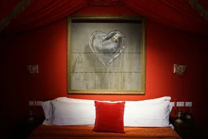 A red bedroom with a wall-mounted artwork featuring a heart with crossbandages, flanked by sconces and a bed with white linen