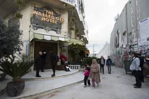 A bustling hotel entrance with concrete walls and glass doors, surrounded by potted plants and people.