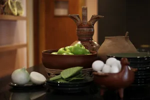 A still life arrangement featuring green apples in a bowl, eggs, sliced onions, and various ceramic bowls and dishes.