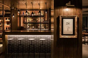 A modern bar interior featuring wooden walls, glass shelves filled with bottles, metal stools, and a brick accent wall.