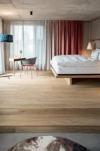 A modern bedroom featuring a wooden floor, a comfortable bed with a white mattress, and a desk area with a chair and a lamp.