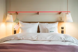 A modern, minimalist bedroom featuring a bed with white pillows and a pink throw blanket, a wooden headboard, and pendant lig