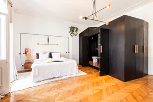 Modern minimalist bedroom with white bed and wooden parquet flooring, featuring a black wardrobe and a plant hanging on the w