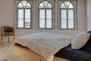 A minimalist bedroom with a white bedframe and blue bedding, featuring arch-shaped windows with white pleated shutters.