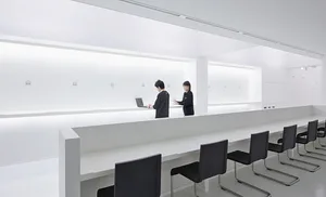 A minimalist office space with white walls, featuring open seating areas and a person working on a laptop.