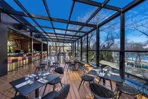 An outdoor restaurant terrace with modern design, featuring wooden flooring, glass ceiling, and metal furniture.