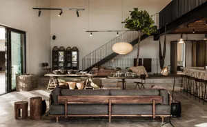 Modern, minimalist living room with concrete and wood elements, featuring a large sofa, wooden stools, and a suspended plant.