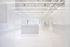 A long, white, minimalist office hallway with glass walls and overhead lighting.