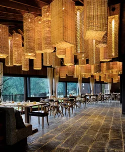 Modern restaurant interior featuring wicker chandeliers and stone flooring, with large windows overlooking a forest.