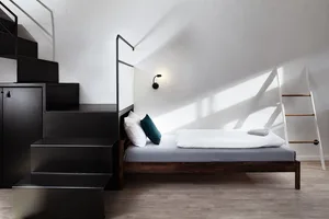 Minimalist bedroom with a wooden full-length bed, white bedding, and a black staircase leading to a cabinet.