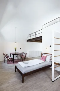 A minimalist bedroom with a single bed, wooden floor, metal ladder, and carpeted area rug.