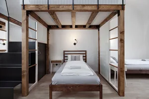 A minimalist bedroom with a single bed on a raised wooden frame, surrounded by transparent glass walls and metal railings.